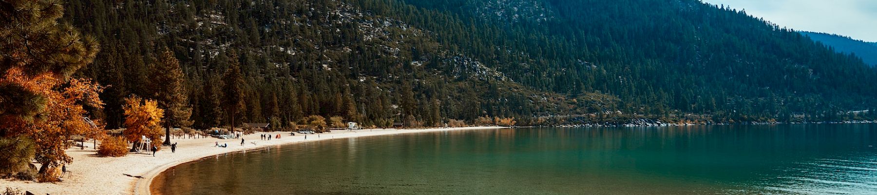 A scenic lake bordered by mountains with a sandy beach, wooden walkway, trees, and calm waters under a partly cloudy sky.