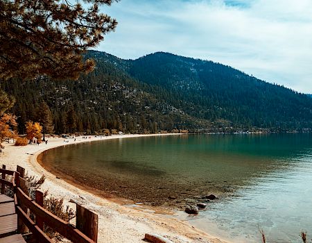 A scenic lake bordered by mountains with a sandy beach, wooden walkway, trees, and calm waters under a partly cloudy sky.