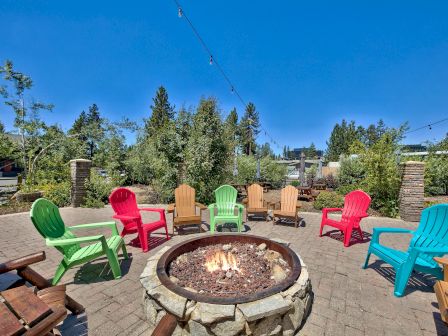 A stone fire pit is circled by colorful chairs in an outdoor patio area with string lights above, surrounded by greenery under a clear blue sky.