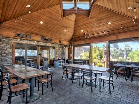 An outdoor dining area with wooden ceiling, multiple tables and chairs, brick floor, and string lights. The walls have large windows and stone decor.