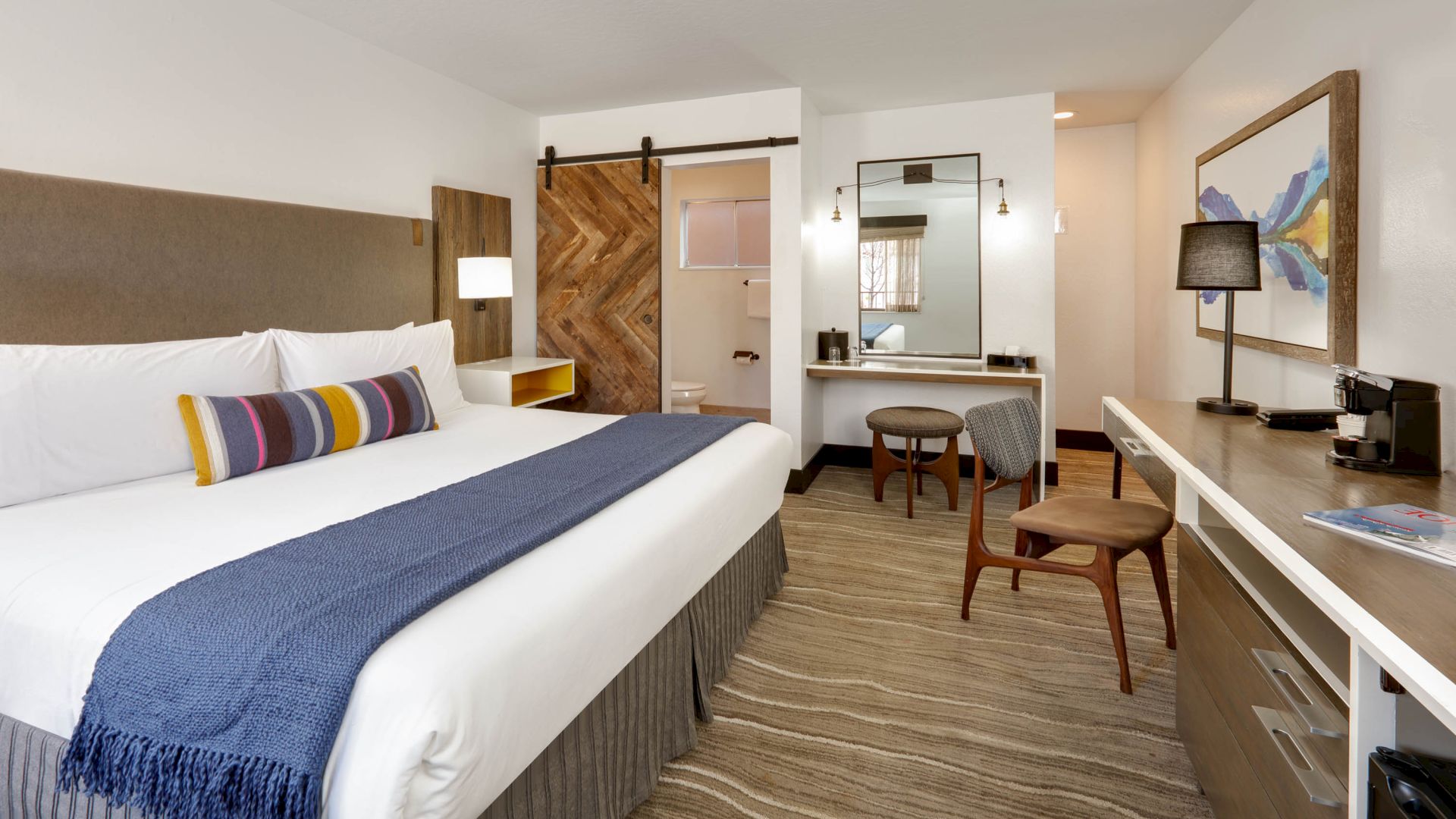 A modern hotel room with a large bed, blue throw, work desk, chair, vanity area, and bathroom with sliding barn door and contemporary decor.