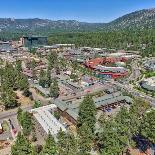 An aerial view of a small town surrounded by a dense forest and mountains, featuring buildings, roads, and parking lots in a scenic landscape.