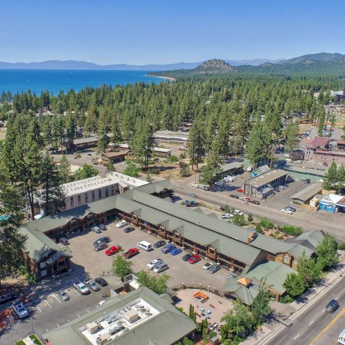 An aerial view of a town with tree-lined streets, buildings, a lake in the background, and mountains in the distance, under a clear blue sky.