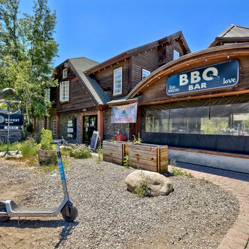 A rustic building houses a BBQ bar with outdoor seating. Nearby, a scooter is parked, and a sign for the adjacent 