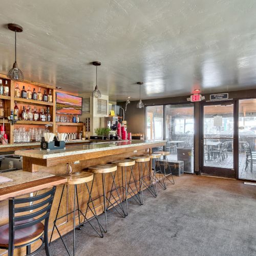 The image shows an empty bar with wooden stools, high tables, a stocked bar, pendant lights, and glass doors leading to an outdoor seating area.