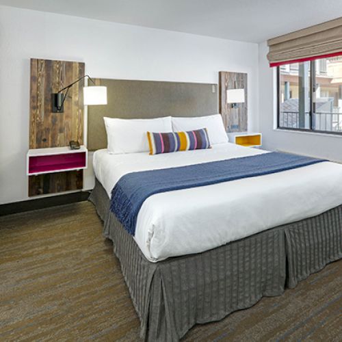 A tidy hotel room with a double bed, colorful pillows, wall lamps, and rustic decor elements. A window and a closed door are in the background.