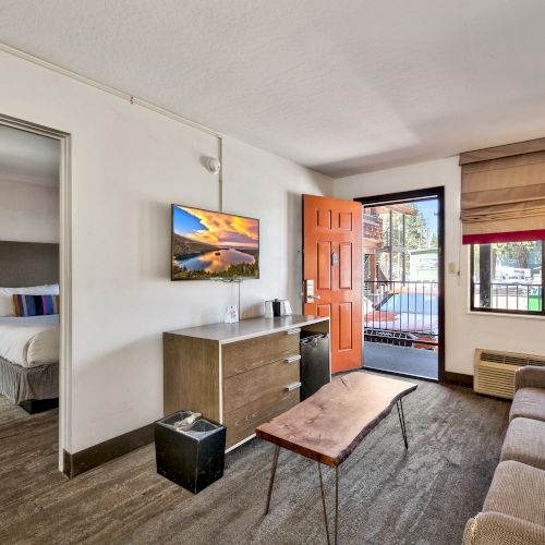 A modern living room with a sofa, wooden table, wall-mounted TV, and open door to a balcony. Connected bedroom visible through a doorway.