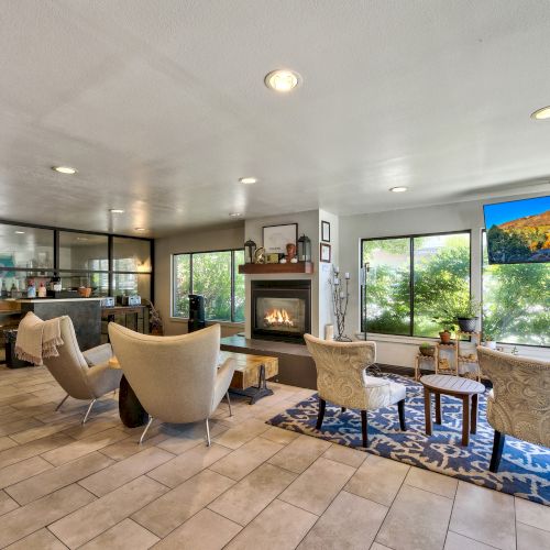 A modern lounge area with cozy seating, a fireplace, large windows, and a blue rug on tiled flooring.