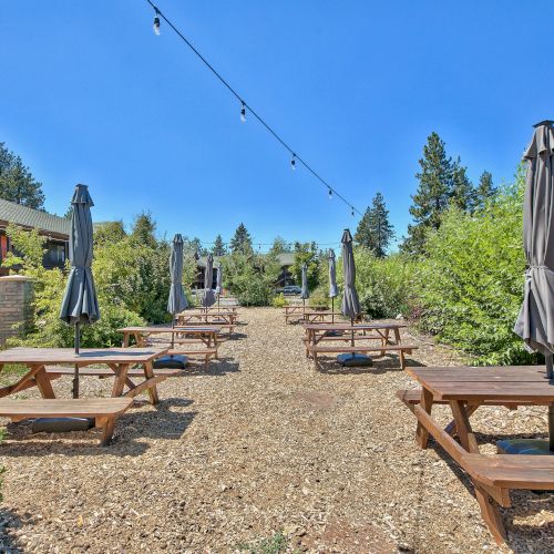 The image shows an outdoor seating area with multiple picnic tables and closed umbrellas, surrounded by trees and a building in the background.