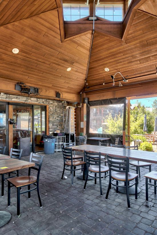 This image shows an outdoor dining area with wooden ceilings, several tables and chairs, and large windows providing a view of greenery outside.