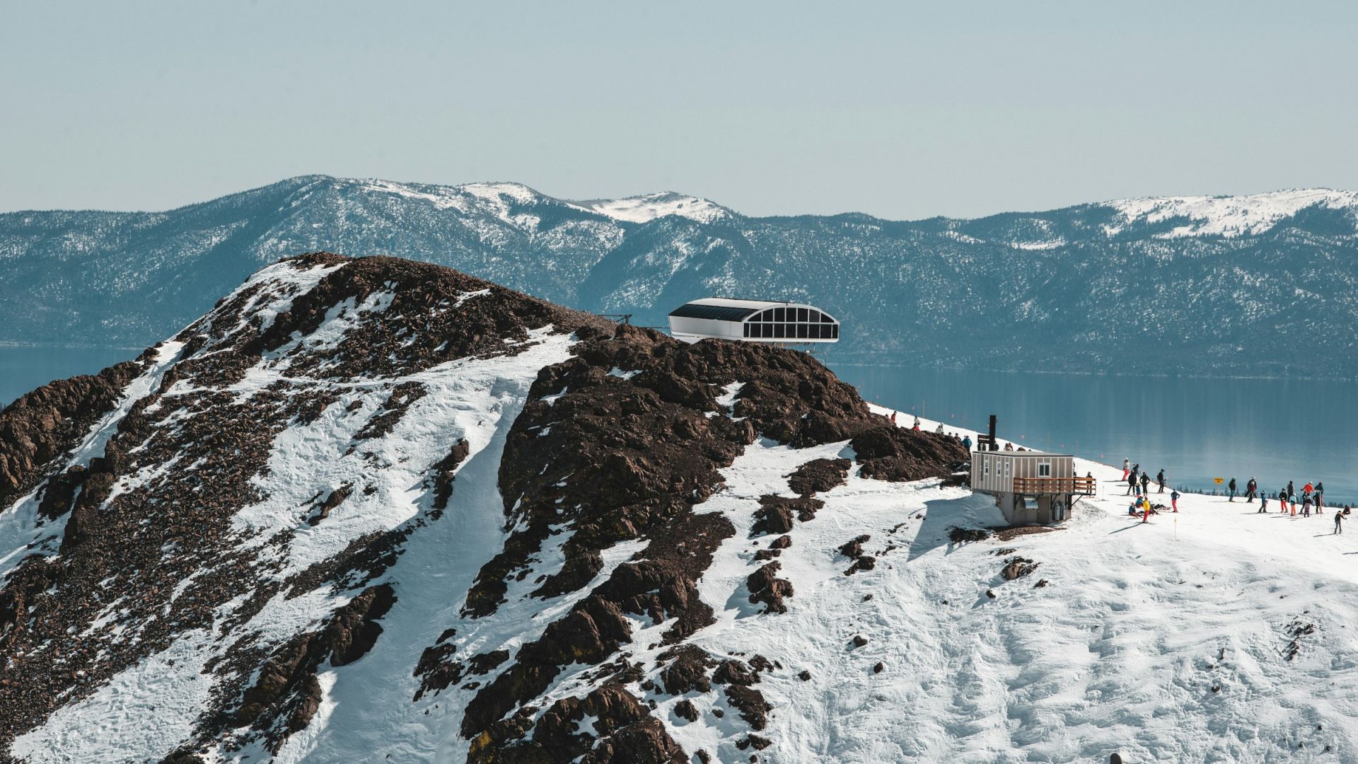 A snowy mountain peak with distant mountains in the background, featuring a modern building structure and people on the slopes.