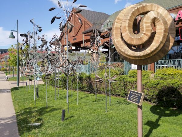 The image shows a sculpture garden with metal wind sculptures on a grassy area, a wooden circular sculpture, and buildings in the background.