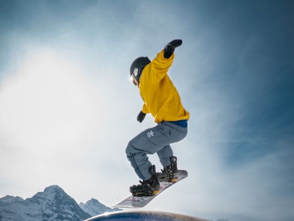 A snowboarder in a yellow jacket is performing a trick on a snowy rail with mountains in the background, under a clear sky.