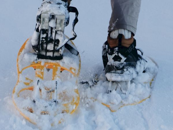 The image shows a person wearing snowshoes while walking on snow-covered ground, with their legs visible from the knee down.