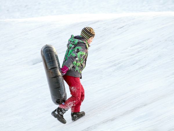 A person in winter clothing carries a black sled or inner tube up a snowy slope, preparing for a downhill ride.