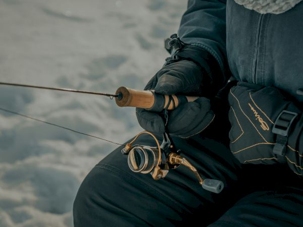 A person in winter gear holds a fishing rod, appearing to be ice fishing in a snowy environment.