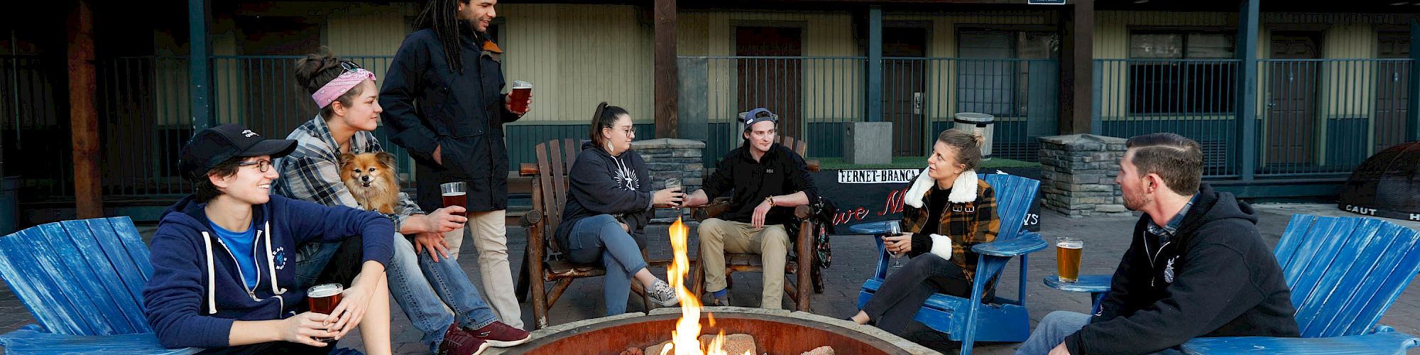 A group of people are sitting around a fire pit, conversing and enjoying drinks. They are seated in blue chairs in front of a building.