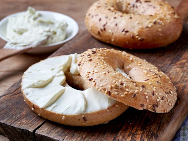 Two sesame bagels on a wooden board, one with cream cheese spread, next to a bowl of extra cream cheese and a wooden knife.
