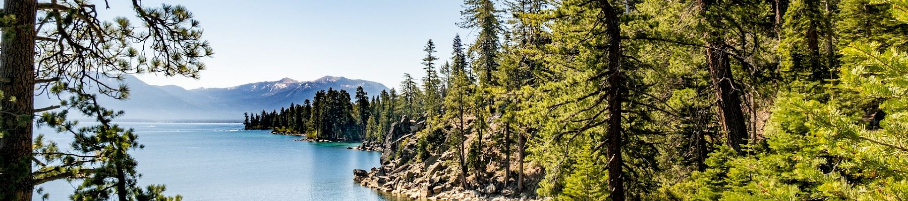 A scenic landscape featuring a clear blue lake surrounded by trees and mountains under a bright blue sky. The water is calm and reflective.