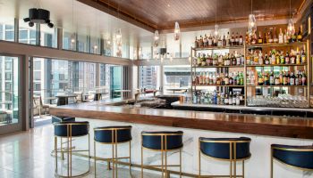 This image shows a modern bar with blue and gold stools, a wooden countertop, and shelves filled with bottles, located in a high-rise building.