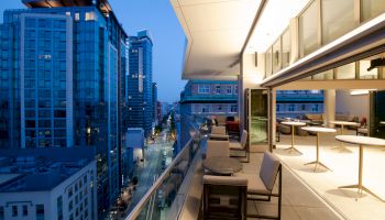 A modern, urban cityscape at dusk, featuring a patio with tables and chairs on a high-rise building, overlooking tall glass skyscrapers.