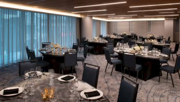 A banquet hall is set up with round tables, each adorned with tableware, black napkins, and floral centerpieces. Blue curtains cover the windows.