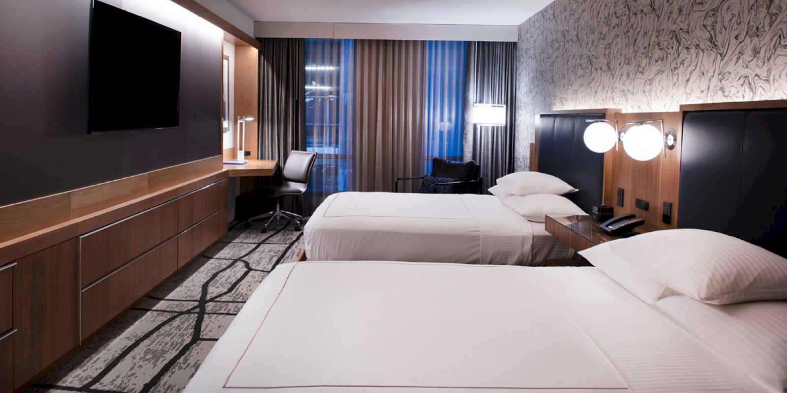 The image shows a modern hotel room with two neatly made beds, a large TV, a desk with a chair, and stylish lamps on the bedside tables.