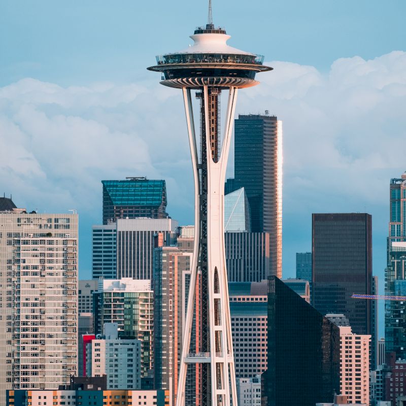 The image shows a cityscape with the Space Needle tower prominently in the center, surrounded by modern buildings, under a partly cloudy sky.