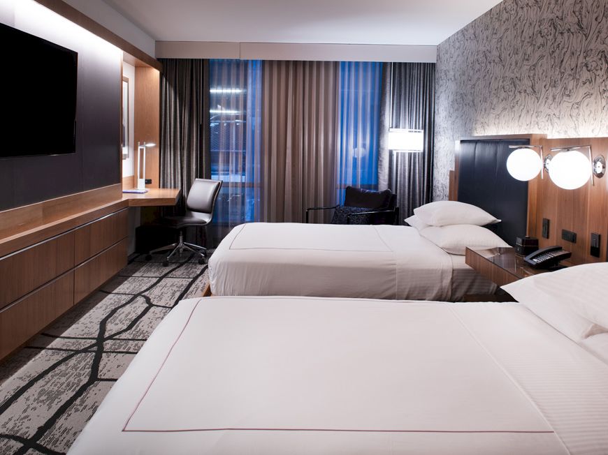 The image shows a modern hotel room with two beds, a large TV, a desk and chair, bedside lamps, and large windows with curtains ending the sentence.