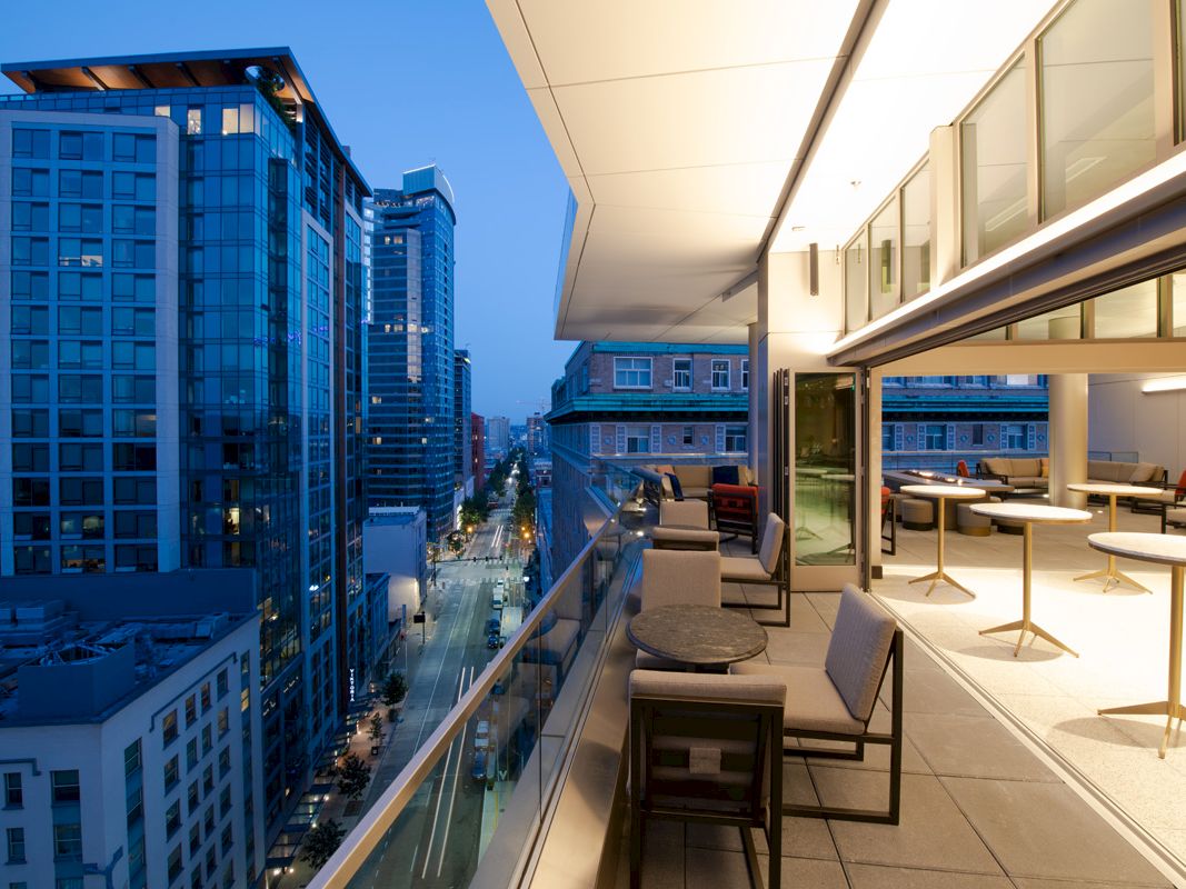 A modern cityscape with high-rise buildings, seen from a balcony with tables and chairs, in the evening light.