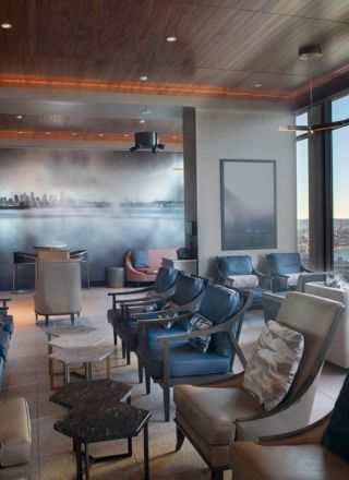 A modern lounge area with comfortable seating, large windows offering a water view, and stylish decor, including a wall mural depicting a cityscape.