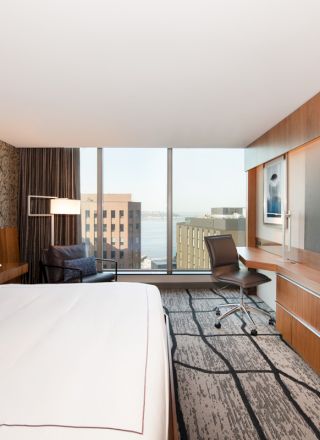 A modern hotel room with a bed, desk, chair, large window view of buildings, and a flat-screen TV mounted on the wall.