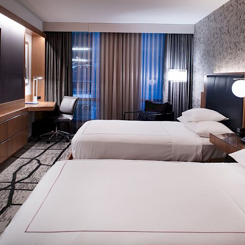 This image shows a modern hotel room with two neatly made beds, a TV on the wall, a work desk by a window, and contemporary decor.