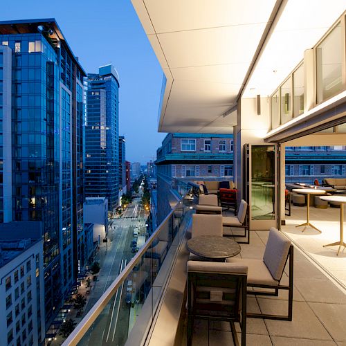 A modern cityscape at dusk with high-rise buildings, viewed from a luxurious balcony featuring seating and tables, overlooking a street below.