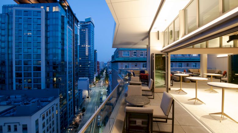 A modern cityscape with tall buildings at dusk, a rooftop terrace with tables and chairs, and indoor seating visible through large windows.