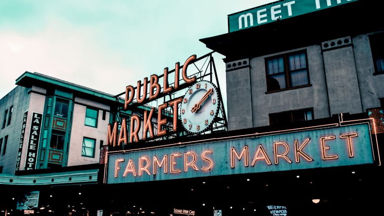 A public market sign with a clock and a "Farmers Market" neon sign in front of buildings. The scene appears lively and urban, suggesting an active market.