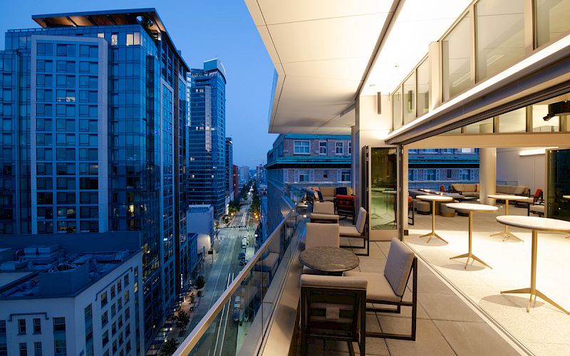 A modern cityscape with high-rise buildings at dusk, featuring a spacious balcony with seating and tables overlooking the street below.